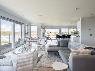 A living room with gray sofas and chairs near large windows overlooking the ocean at Seascape in Polzeath