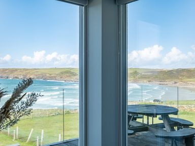 A view of the ocean and coastline through two large windows with a wooden picnic table outside on a deck at Seascape Polzeath