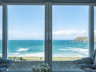 A view of the ocean and coastline through large glass doors with a wooden deck and seating inside at Seascape Polzeath