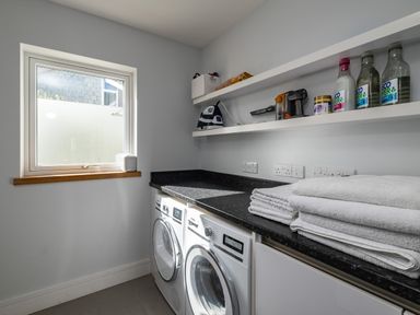 A laundry room with stacked towels washing machine dryer and shelves with cleaning supplies and an iron at Seascape in Polzeath