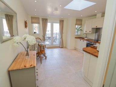 A kitchen with white cabinets wooden countertops a dining table with chairs flowers on a dresser and glass doors leading outside at Bumble Cottage in Tysoe