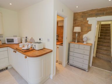 A kitchen area with wooden countertops microwave toaster cups plates and a staircase next to a chest of drawers with a lamp and decor at Bumble Cottage in Tysoe