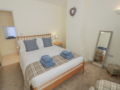 A bedroom with a double bed with blue towels and cushions a side table with lamp a full length mirror a chair and a small footstool at Bumble Cottage in Tysoe