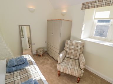 A bedroom with a plaid armchair a matching footstool a wardrobe a large mirror and a bed with folded towels at Bumble Cottage in Tysoe