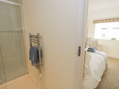 A view from a bathroom with a shower and towel rack into a bedroom with a bed and armchair at Bumble Cottage in Tysoe