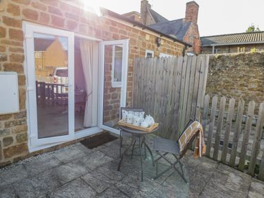 A small outdoor patio area with a table and two chairs next to a stone wall and a glass door at Bumble Cottage in Tysoe