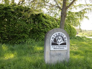 A stone sign with a sheep illustration and text indicating the Cotswolds area of outstanding natural beauty in a grassy area with trees and bushes at Bumble Cottage in Tysoe