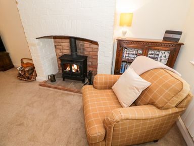 A living room with a wood burning stove a wicker basket with firewood a patterned armchair and a wooden cabinet with a lamp and books at Bumble Cottage in Tysoe