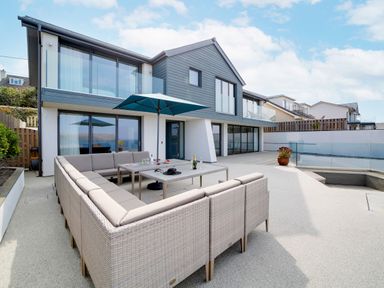 An outdoor patio area with a large cushioned seating arrangement under an umbrella in front of a modern two-story house with glass balcony at Megizzy in Polzeath