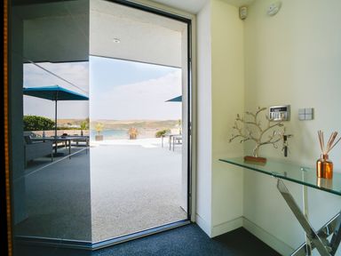 An open doorway leading to a patio with outdoor furniture and umbrellas overlooking a coastal landscape at Megizzy in Polzeath