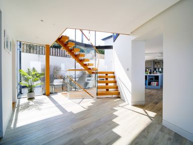 An open hallway with wooden stairs and glass railing next to a small outdoor seating area and a kitchen in the background at Megizzy in Polzeath