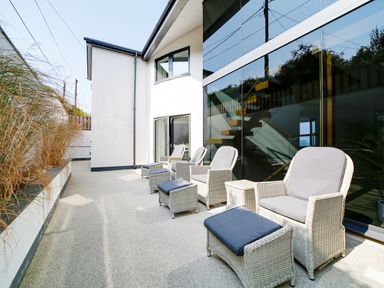 An outdoor patio with wicker chairs and footstools next to a modern building with large glass windows at Megizzy in Polzeath