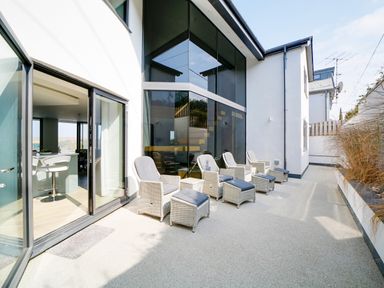 A patio with six wicker chairs and footrests outside a modern house with large glass windows at Megizzy in Polzeath