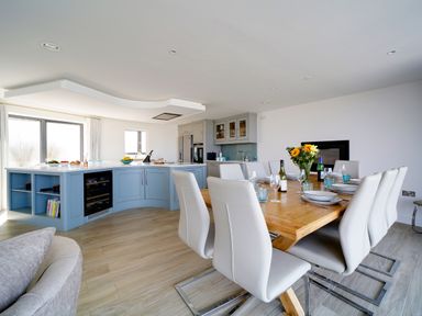 A dining area with a wooden table set for six and a kitchen with blue cabinets in an open plan room at Megizzy in Polzeath