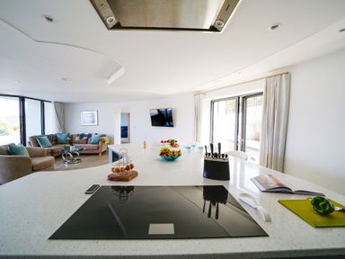 A kitchen island with a cooktop knife block and fruit bowls overlooking a living area with sofas and a wall-mounted TV at Megizzy in Polzeath