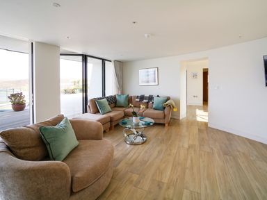 A living room with a brown armchair and sofa with green cushions a glass coffee table and large windows at Megizzy in Polzeath