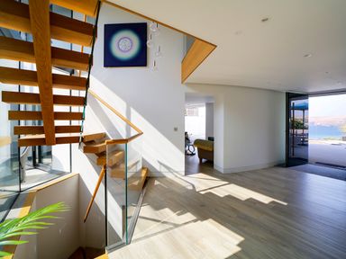 A stairway with wooden steps and glass railing in a hallway with light wood flooring and an open door showing an outdoor view at Megizzy Polzeath