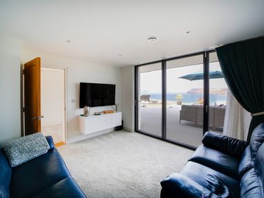 A living room with blue sofas a wall-mounted TV and large sliding glass doors opening to an outdoor patio with a sea view at Megizzy in Polzeath
