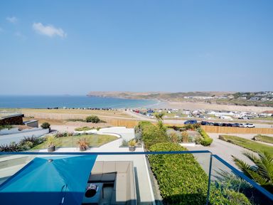 A coastal view with a swimming pool patio and seating area overlooking a beach and parked cars at Megizzy in Polzeath