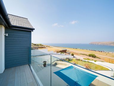 A balcony with glass railing overlooking a garden with plants a blue umbrella and the sea at Megizzy in Polzeath