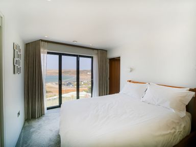 A bedroom with a large bed and white linens with glass doors showing a coastal view at Megizzy in Polzeath