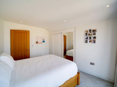 A bedroom with a wooden bed frame and white bedding next to a wooden door and white sliding closet doors with a mirror at Megizzy in Polzeath