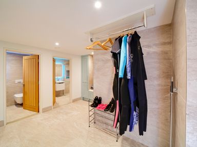 A spacious room with hanging wetsuits on a rack and open doors leading to a bathroom and toilet at Megizzy in Polzeath