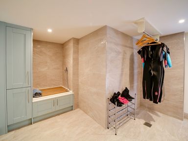 A room with a built-in bench, storage cabinets, a shoe rack, and wetsuits hanging on a rod at Megizzy in Polzeath