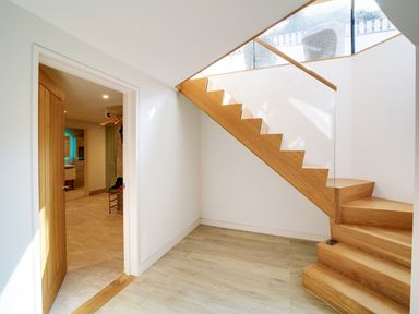 A wooden staircase with glass railing and an open wooden door showing a hallway with beige tiled floor at Megizzy in Polzeath