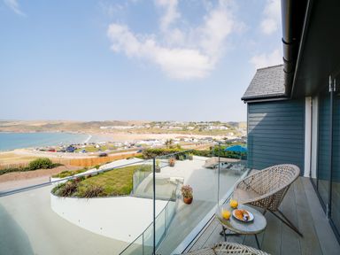 A balcony with two wicker chairs and a small table with juice and croissants overlooking a patio and coastal view at Megizzy in Polzeath