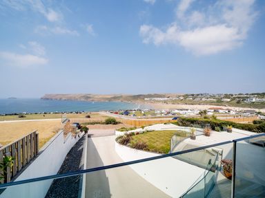 A view of a coastal landscape with a curved garden patio and glass railing overlooking a beach and houses at Megizzy in Polzeath