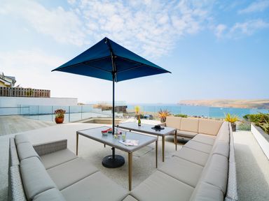 An outdoor seating area with beige cushioned sofas around two tables and a blue umbrella with a sea view at Megizzy in Polzeath