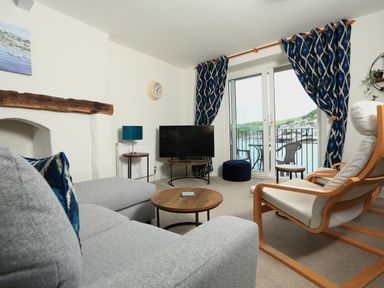 A living room with a gray sofa a wooden armchair a round wooden coffee table a TV on a stand blue patterned curtains and a balcony with chairs overlooking water at Ferry View in Dartmouth