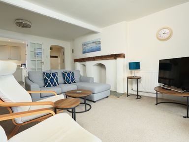 A living room with a gray sofa and blue pillows two white chairs two round wooden tables a TV on a stand a wall clock and a painting above a wooden beam at Ferry View in Dartmouth