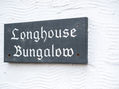 A black rectangular sign with white gothic text reading Longhouse Bungalow on a white textured wall at Longhouse Bungalow in Rock