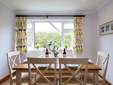 A dining room with a wooden table set for four with white chairs near a window with patterned curtains at Longhouse Bungalow in Rock