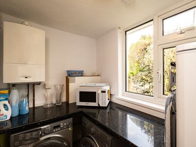 A utility room with a washing machine, dryer, microwave oven, detergent bottles, a water heater, and a window showing outdoor greenery at Longhouse Bungalow in Rock
