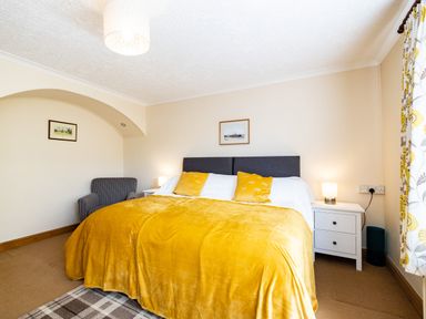 A bedroom with a large bed covered with a yellow blanket and pillows a gray armchair two bedside tables with lamps framed pictures on the walls and patterned curtains at Longhouse Bungalow in Rock
