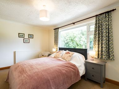 A bedroom with a double bed pink blanket bedside tables with lamps and patterned curtains at Longhouse Bungalow in Rock