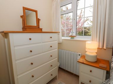 A bedroom corner with a white chest of drawers with a mirror on top a white bedside table with a lamp and a window with curtains overlooking trees at 16 Norfolk Heights in Docking