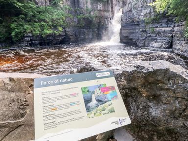 A waterfall flowing into a river with a rock information sign about the force of nature at Foxes Den in Winston near Staindrop