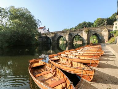 Wooden rowboats tied along the riverbank near a stone arch bridge with trees and buildings nearby at Foxes Den in Winston near Staindrop