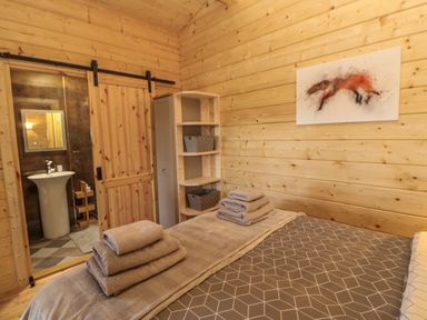 A bedroom with a bed with towels on it a wooden sliding door leading to a bathroom and a corner shelf with baskets and a fox painting on the wall at Foxes Den Winston near Staindrop