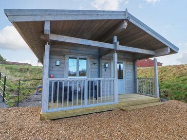 A small wooden cabin with a covered porch and outdoor furniture on a gravel surface at Foxes Den in Winston near Staindrop