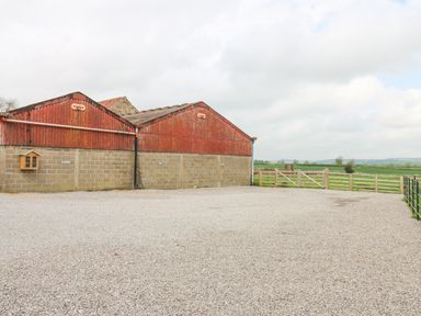 An outdoor area with a gravel ground and two farm buildings with red corrugated roofs near wooden fenced fields at Foxes Den in Winston near Staindrop