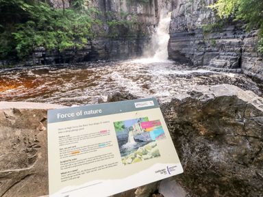 A waterfall with rocks and a sign explaining the force of nature at Otters Holt in Winston near Staindrop