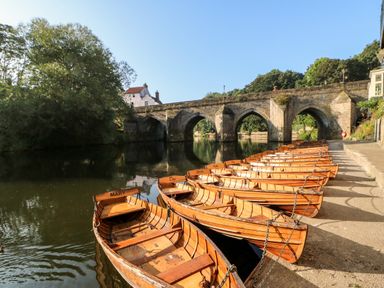 Wooden boats tied along a riverbank with an old stone bridge in the background at Otters Holt Winston near Staindrop
