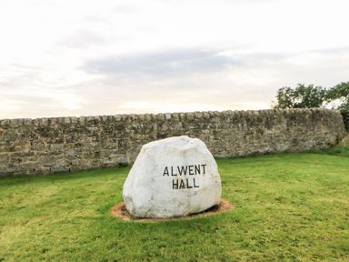 A large white rock with Alwent Hall written on it on grass next to a stone wall