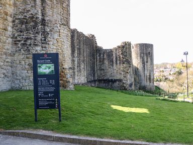 Stone walls and towers of Barnard Castle with a sign displaying information about the site and a grassy area in the foreground at Barnard Castle
