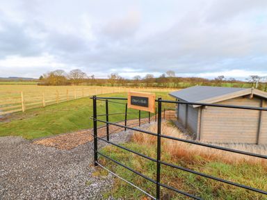A black metal gate with a sign reading Otters Holt next to a gravel path and wooden fence surrounding fields at Otters Holt Winston near Staindrop